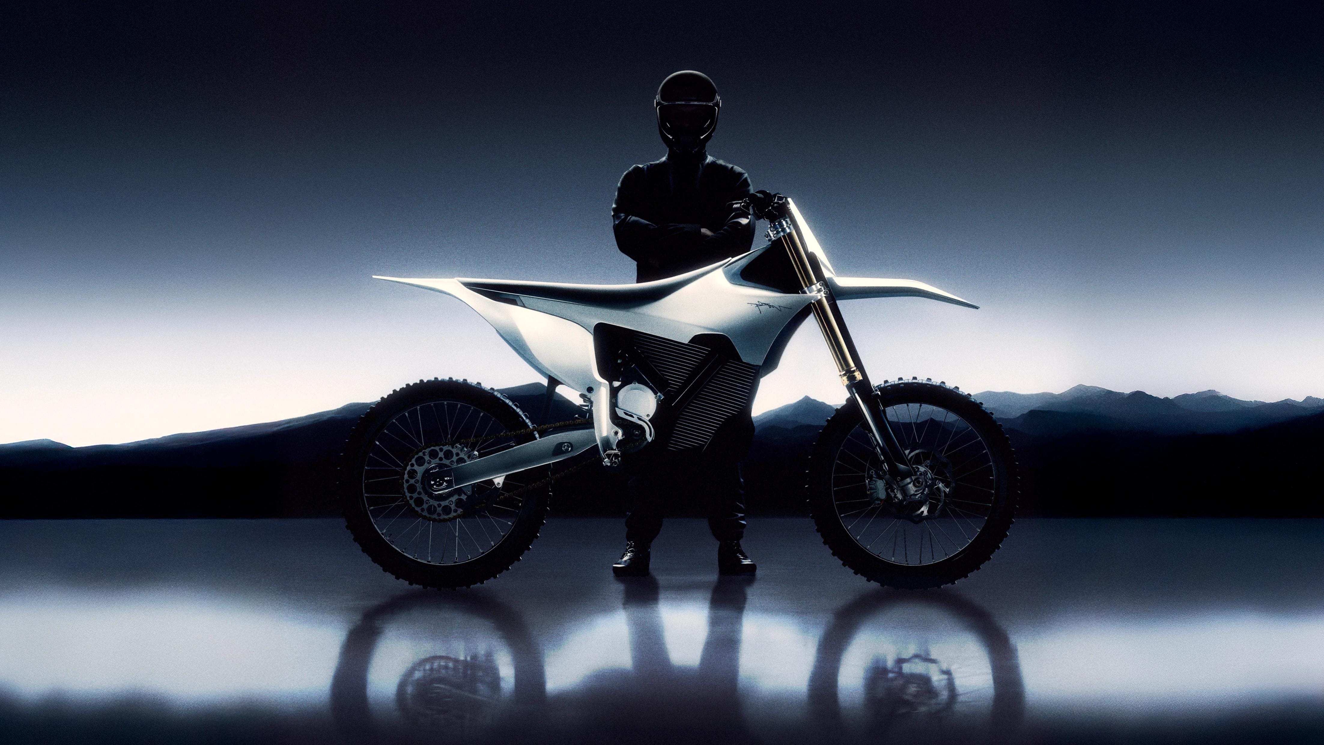 White Stark VARG electric motocross bike and helmeted rider silhouetted against a low blue horizon on a reflective black floor.