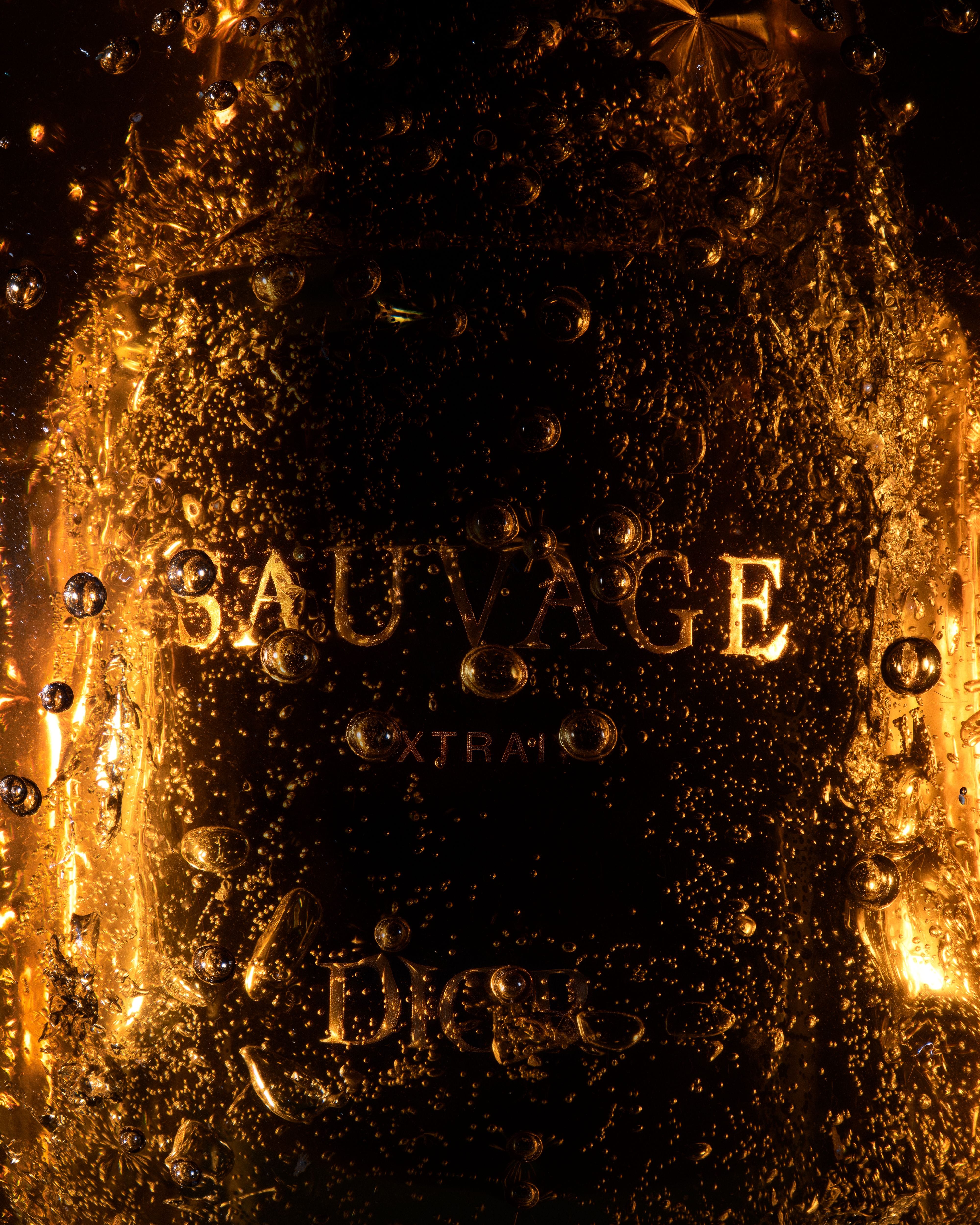 Close view of a Dior Sauvage Extrait bottle almost submerged in dark amber material, with bubbles and reflections across its surface.