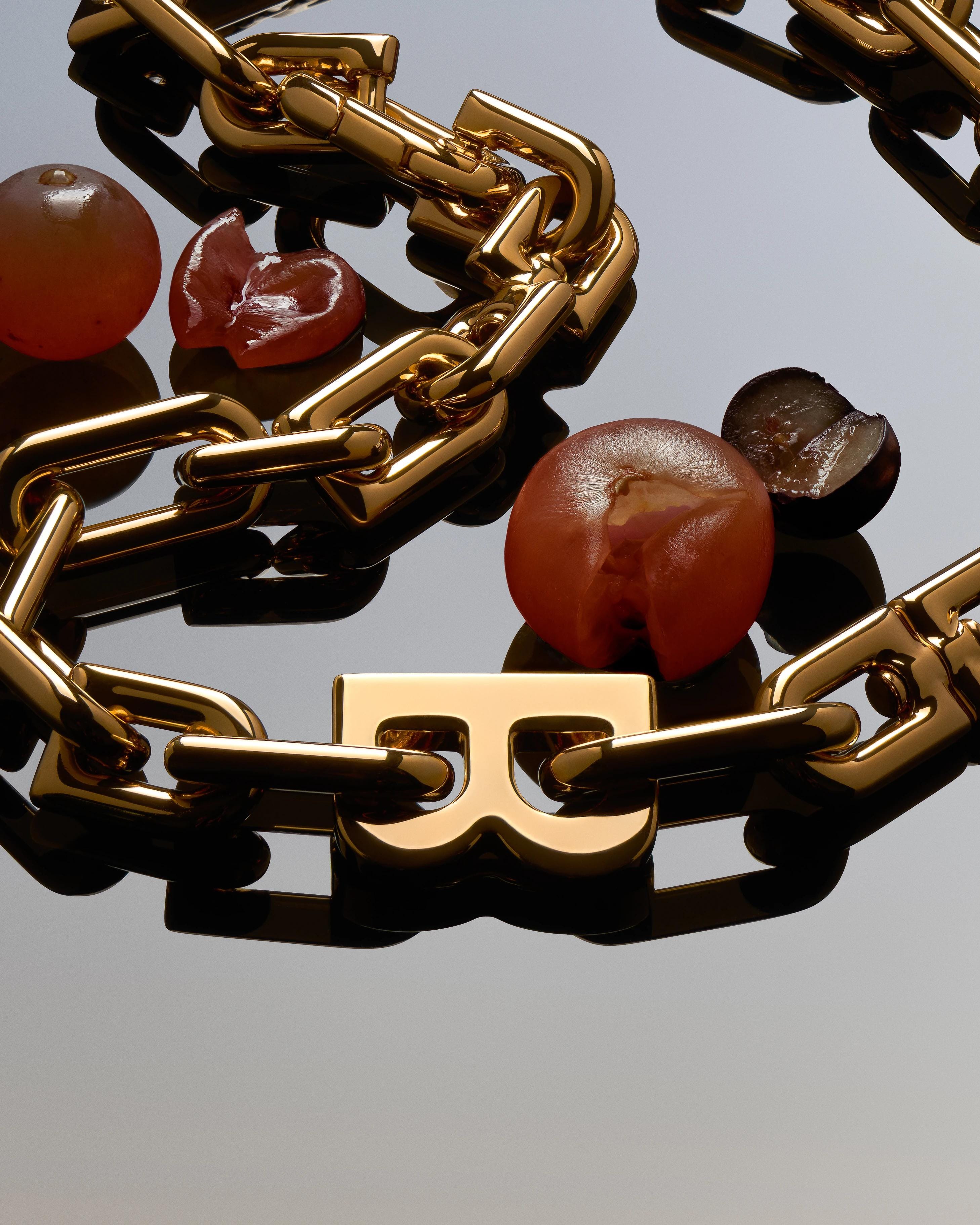 Balenciaga gold-tone B Link necklace arranged with translucent red-brown spheres and glossy fragments on a mirrored surface.