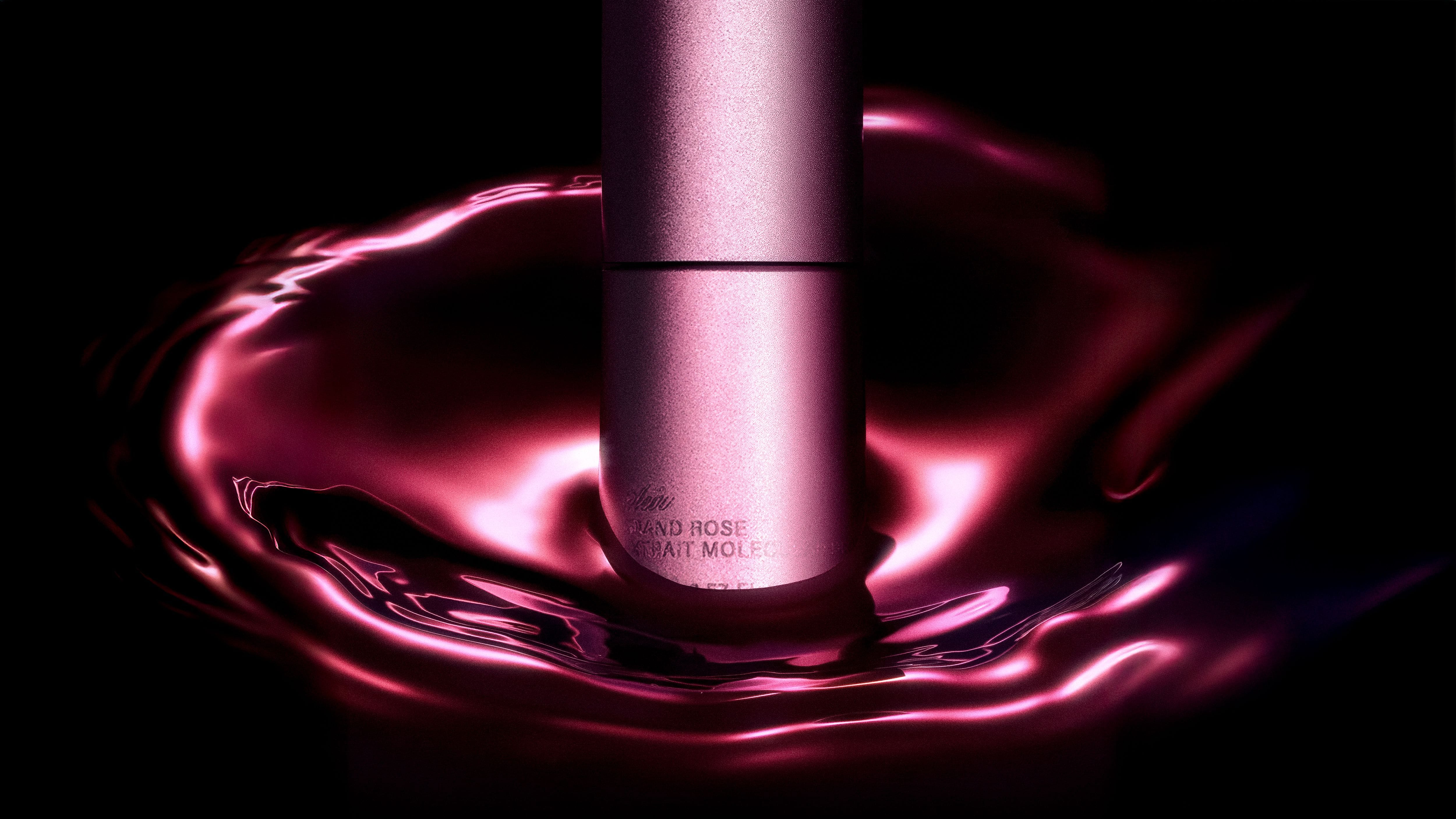 Rose-coloured Aeir fragrance atomiser standing in a glossy magenta liquid ring against black.