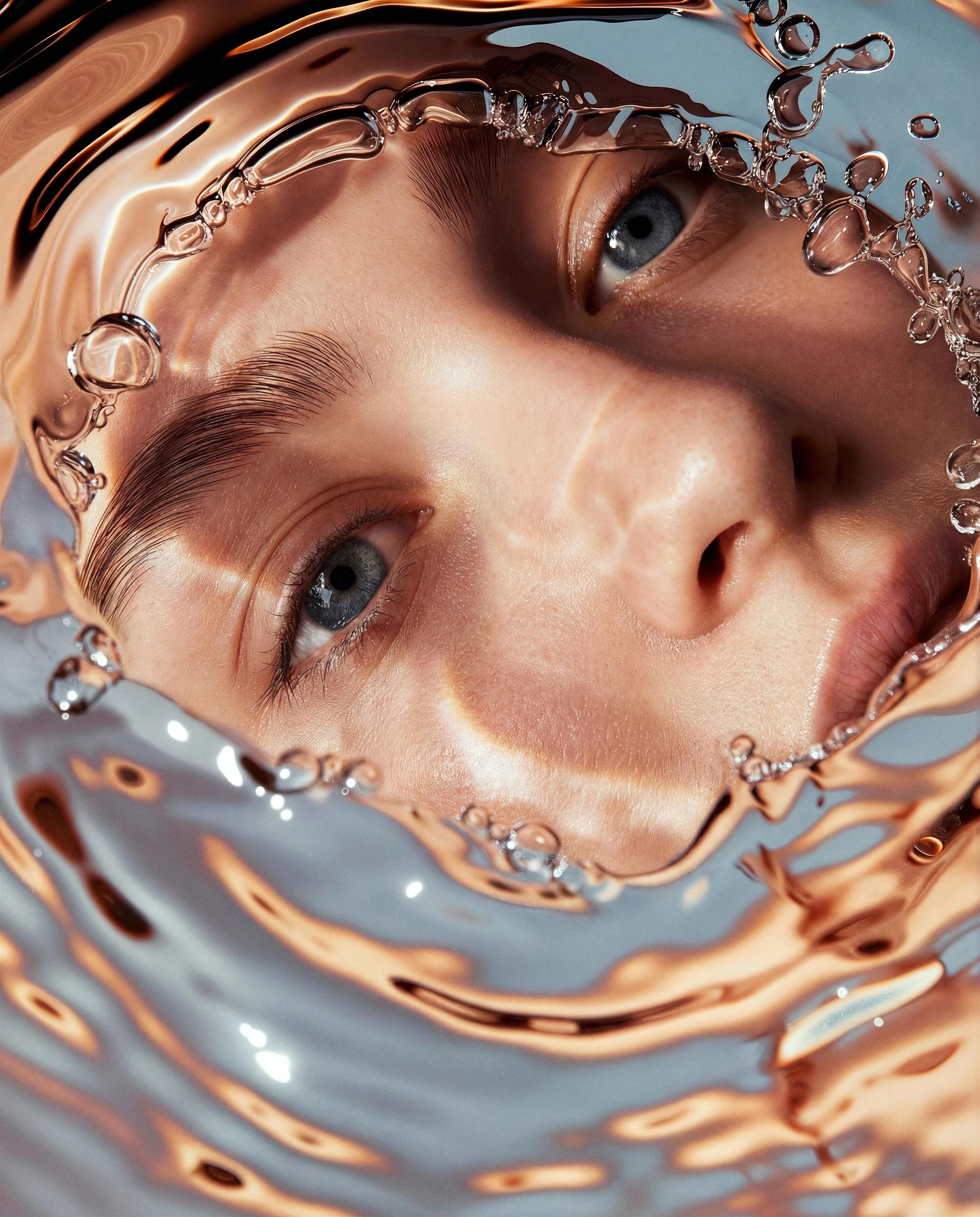 Face distorted through rippling reflective liquid in cool silver-blue light.