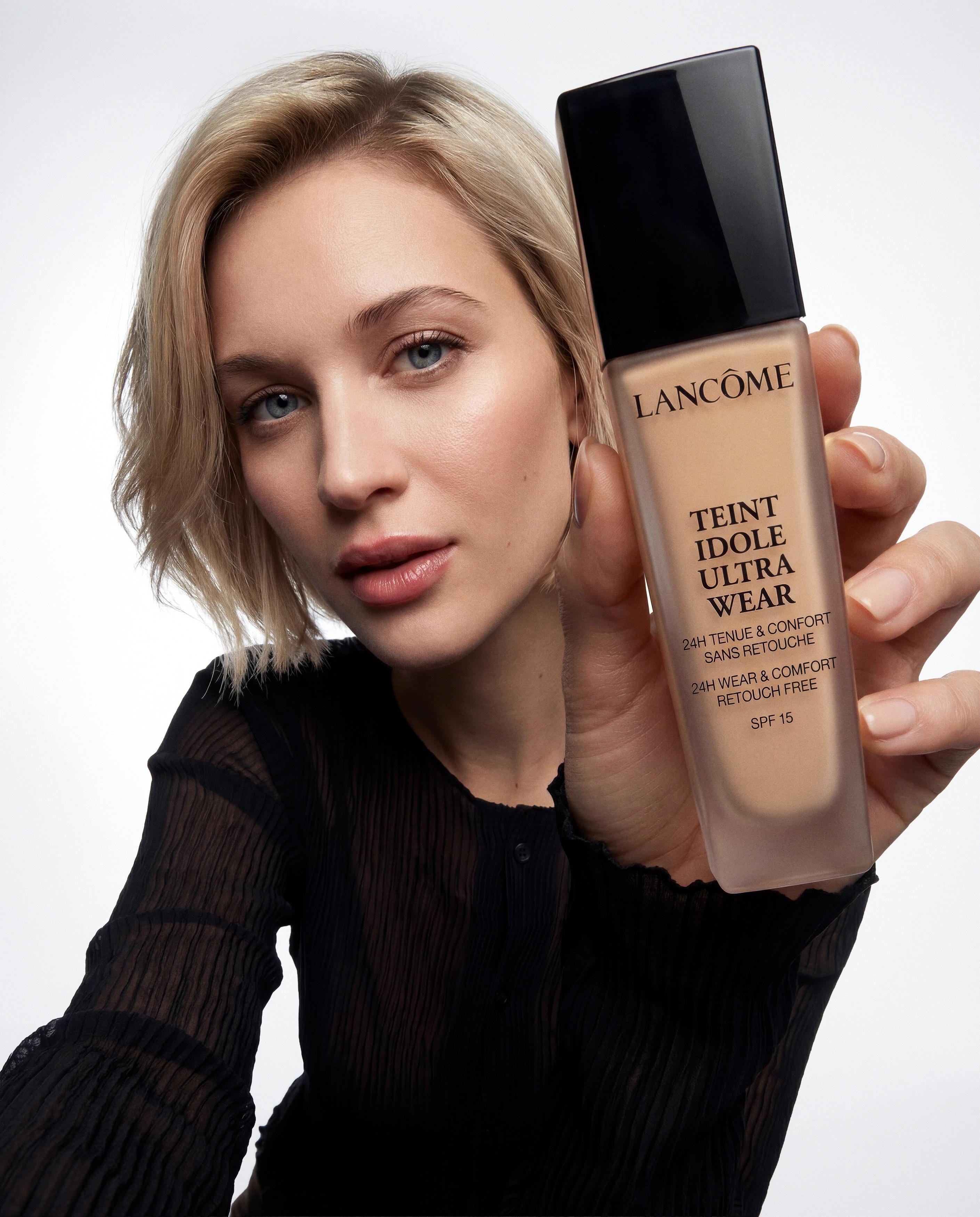 Blonde woman in black holding a Lancôme Teint Idole Ultra Wear foundation bottle towards the camera.