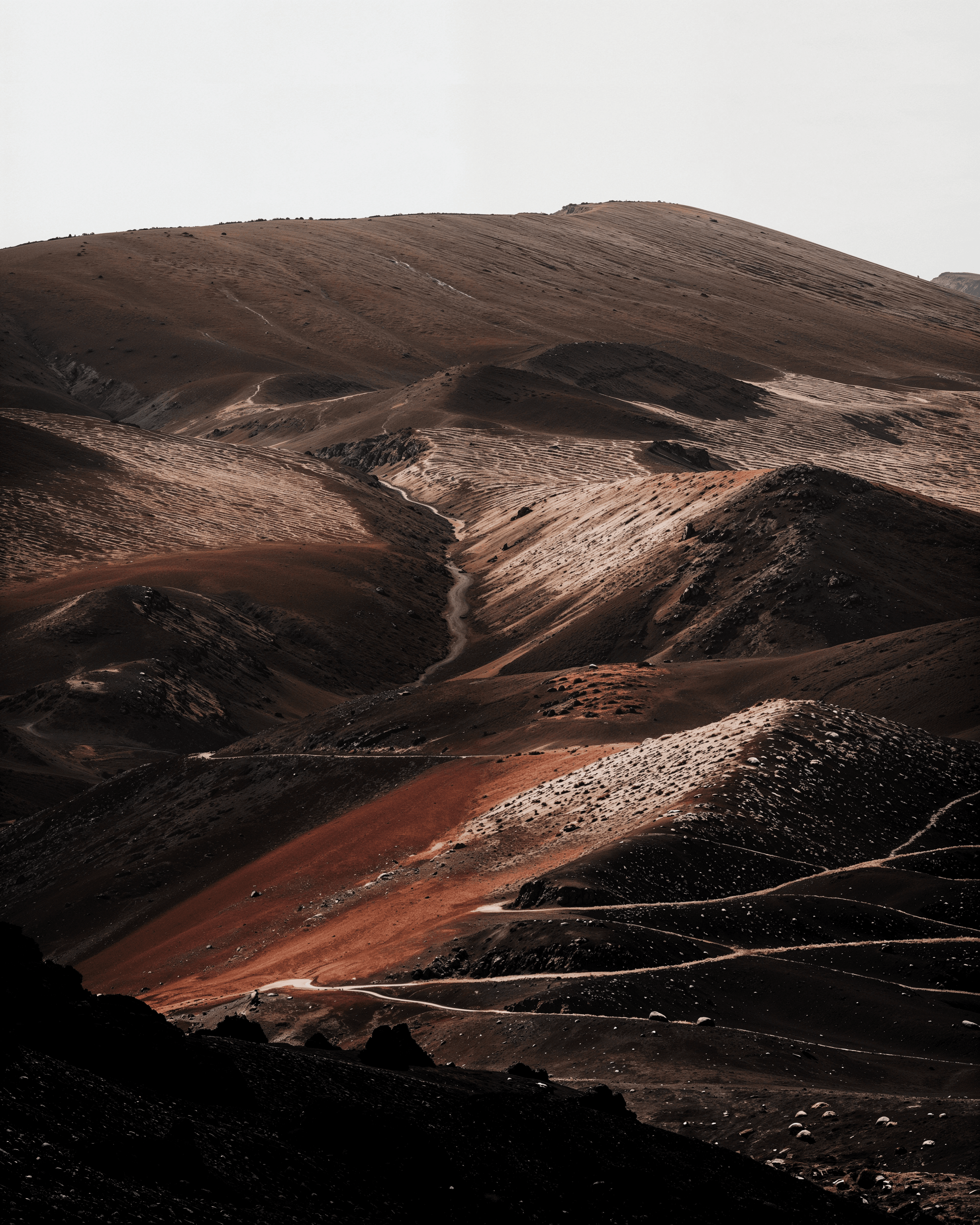Layered mineral-red volcanic ridges crossed by a pale path beneath a flat grey sky.