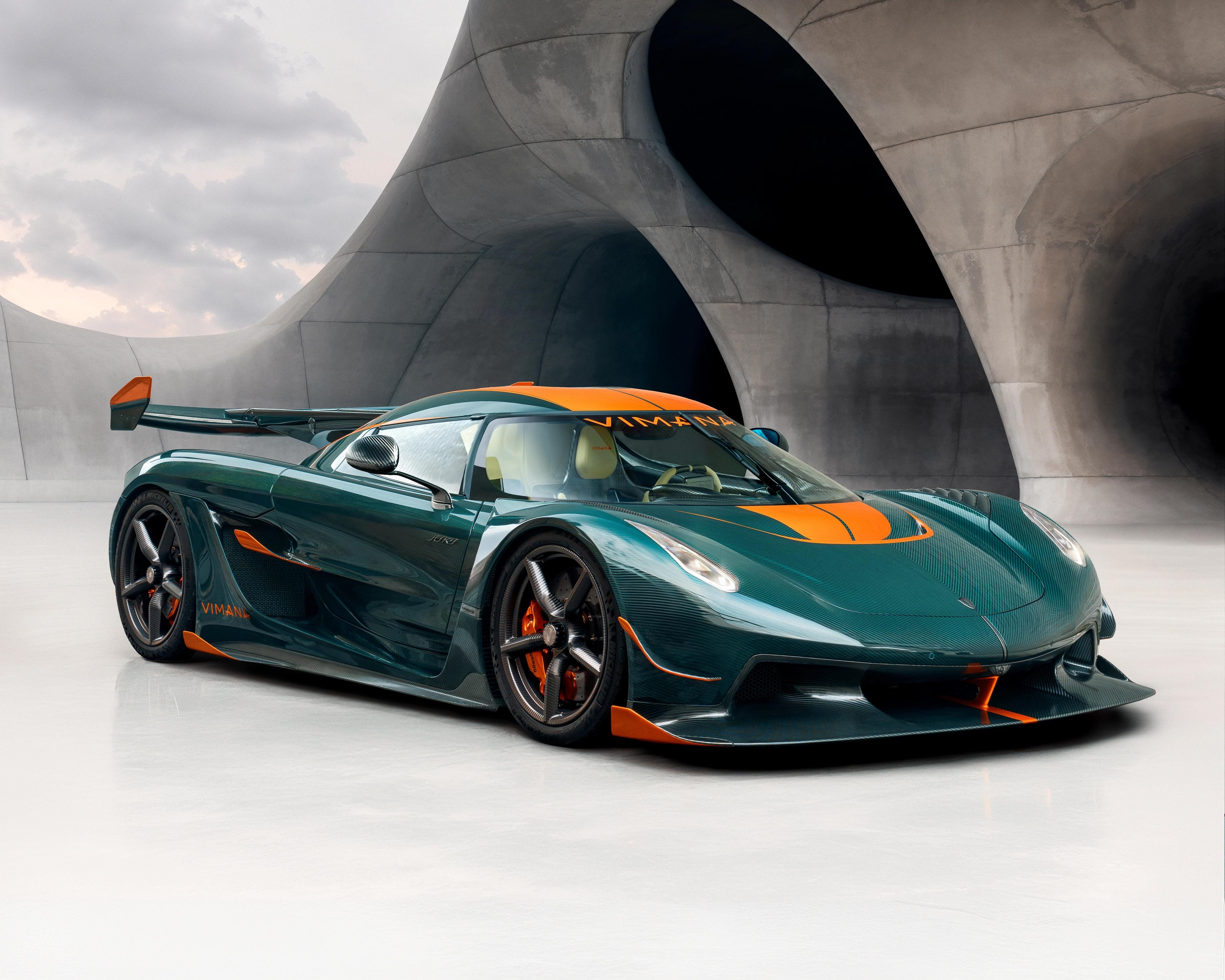 Deep green Koenigsegg Jesko with orange detailing parked in front of sweeping concrete architecture.