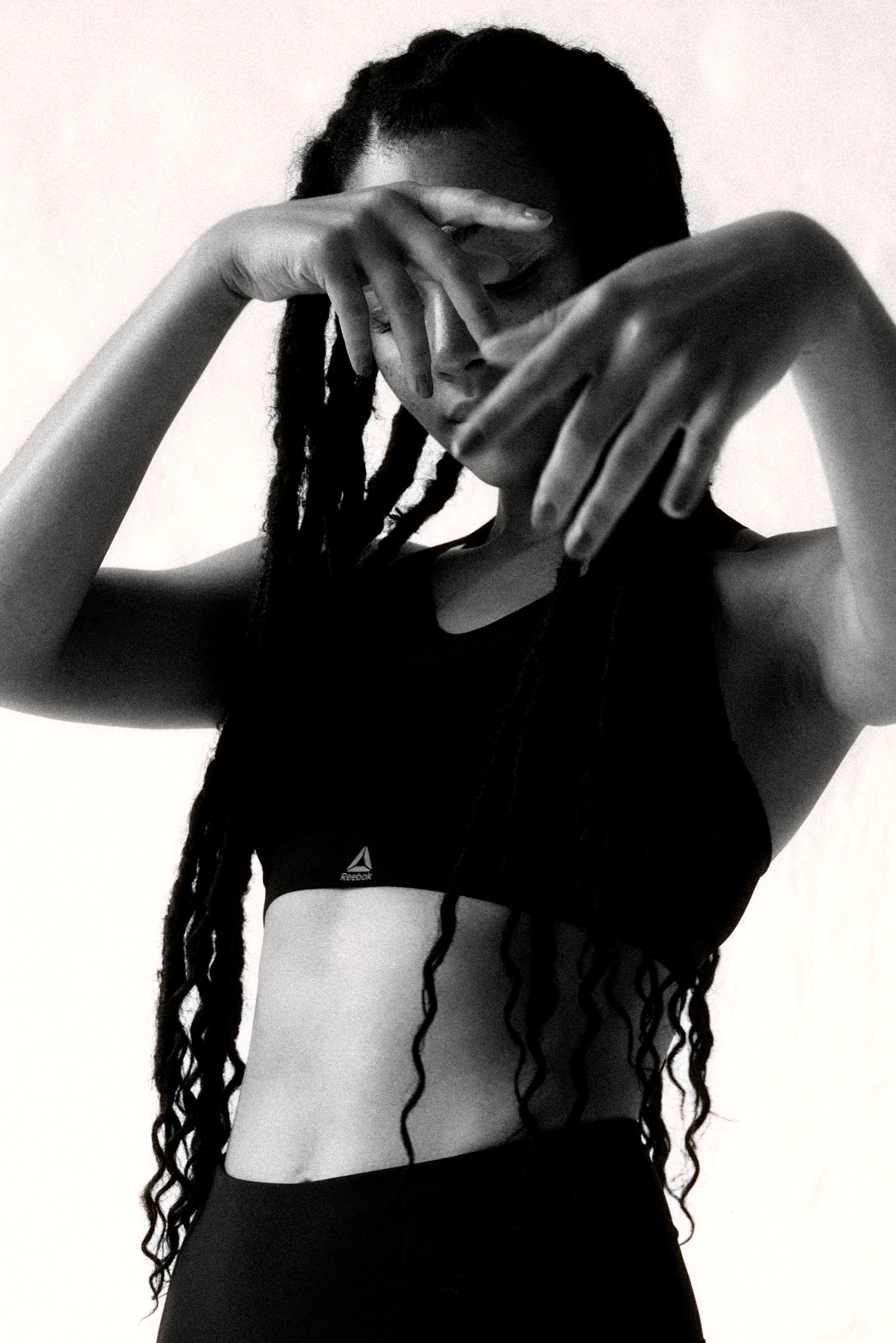 Grainy black-and-white portrait of Julia Dalia wearing the Reebok PureMove Bra, her braided hair and raised hands framing her face.