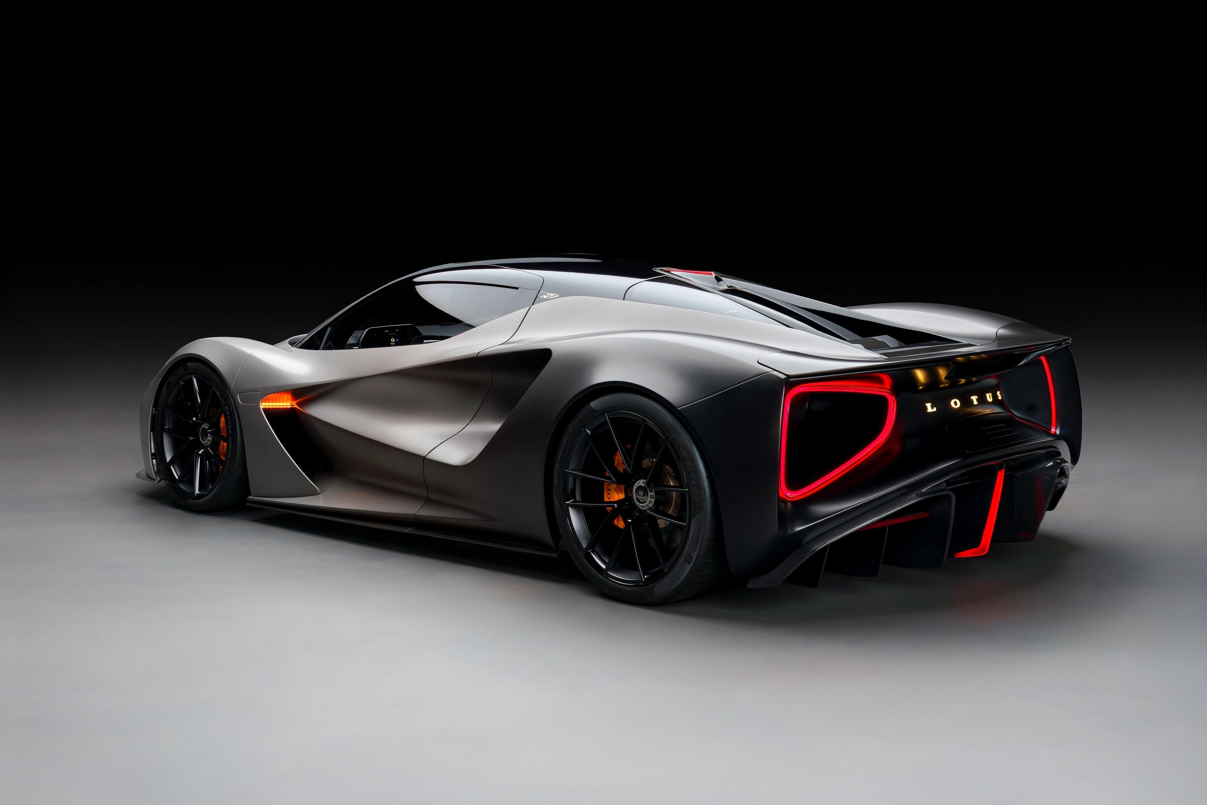 Rear three-quarter view of a graphite Lotus Evija in a dark studio, with its red rear lights illuminated.