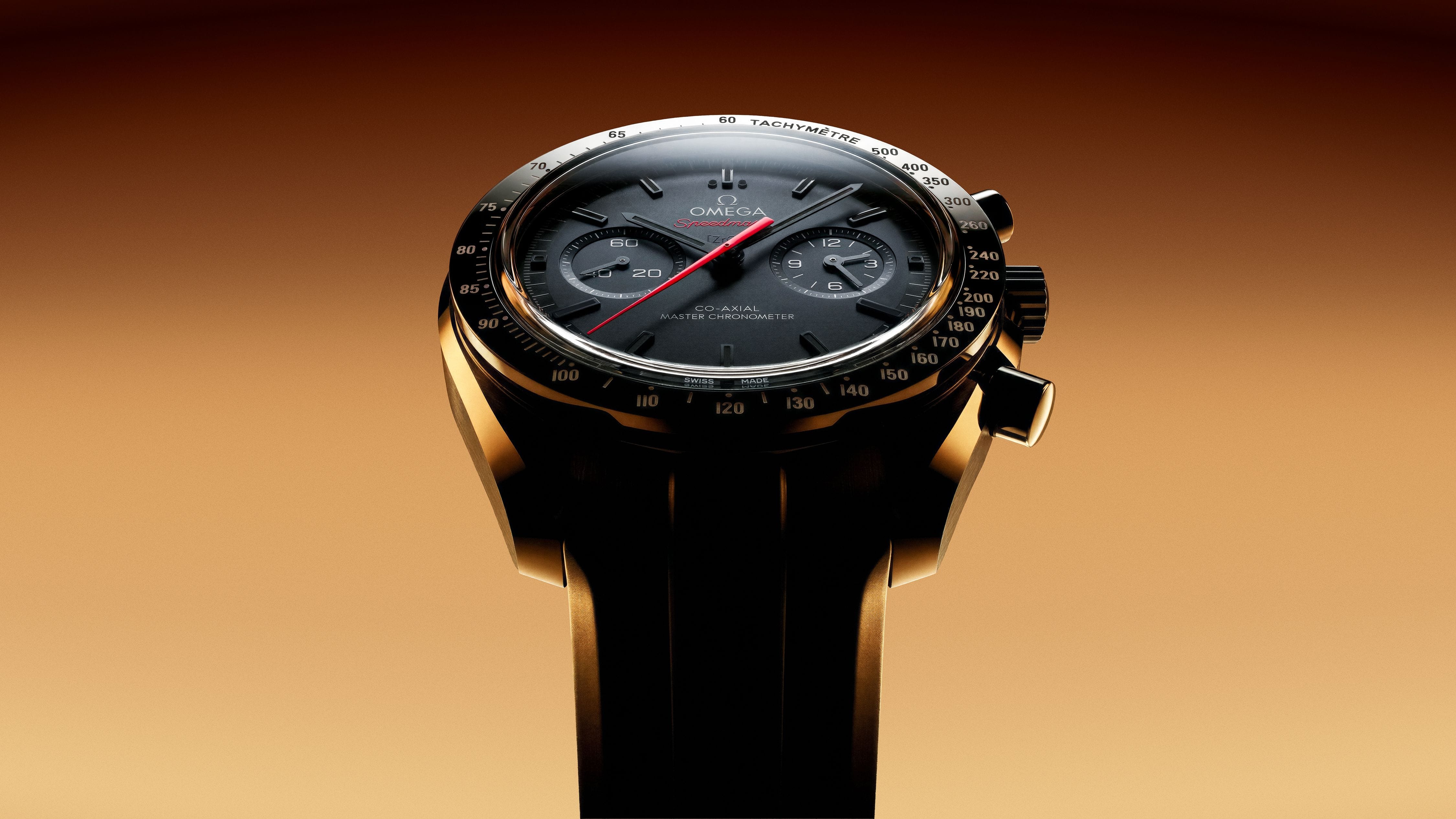 Black Omega Speedmaster rising against amber light, its red chronograph hand and sculpted case held in sharp relief.