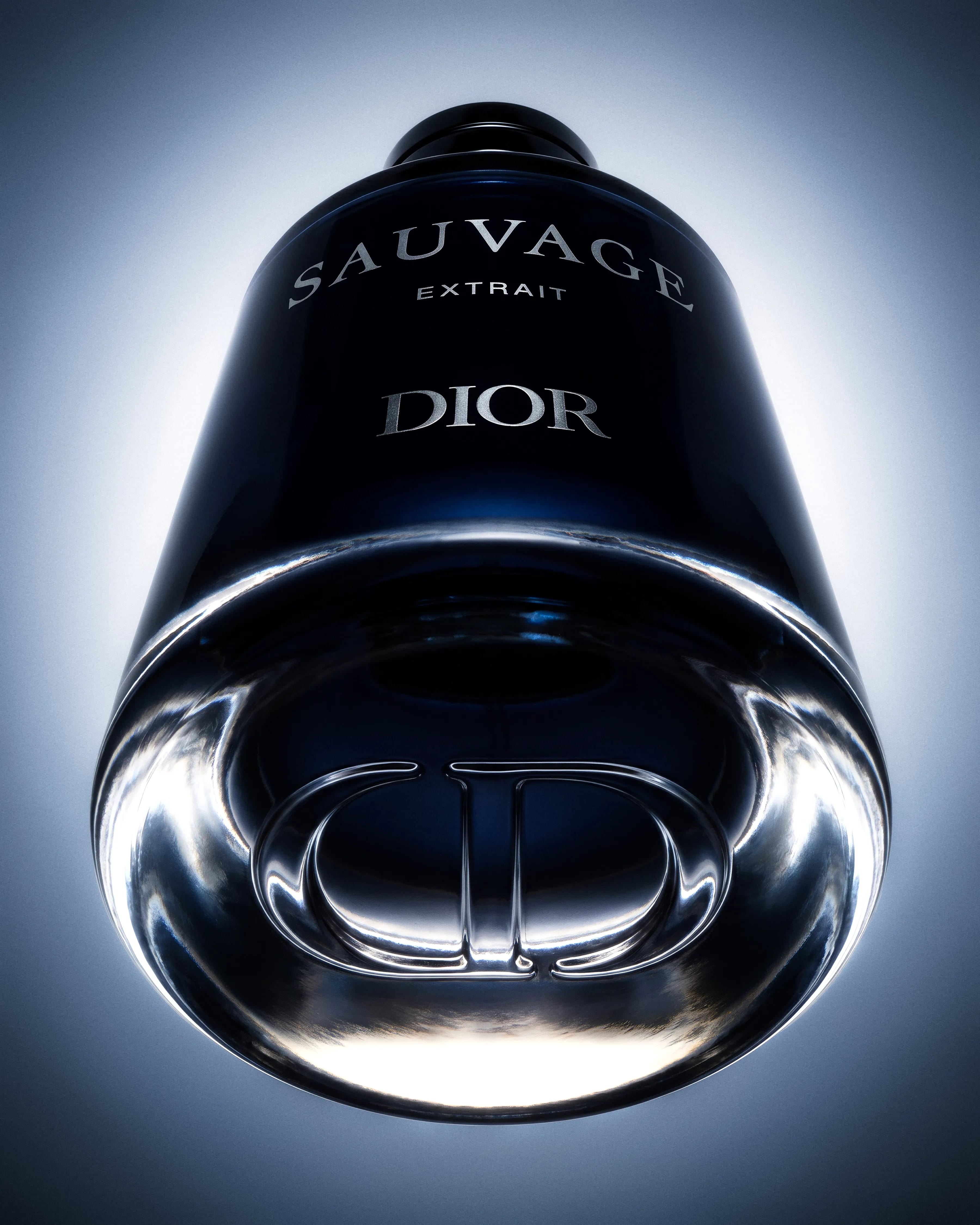 Extreme low-angle photograph of the Dior Sauvage Extrait bottle, enlarging the engraved CD monogram in its clear glass base against cold blue-white light.