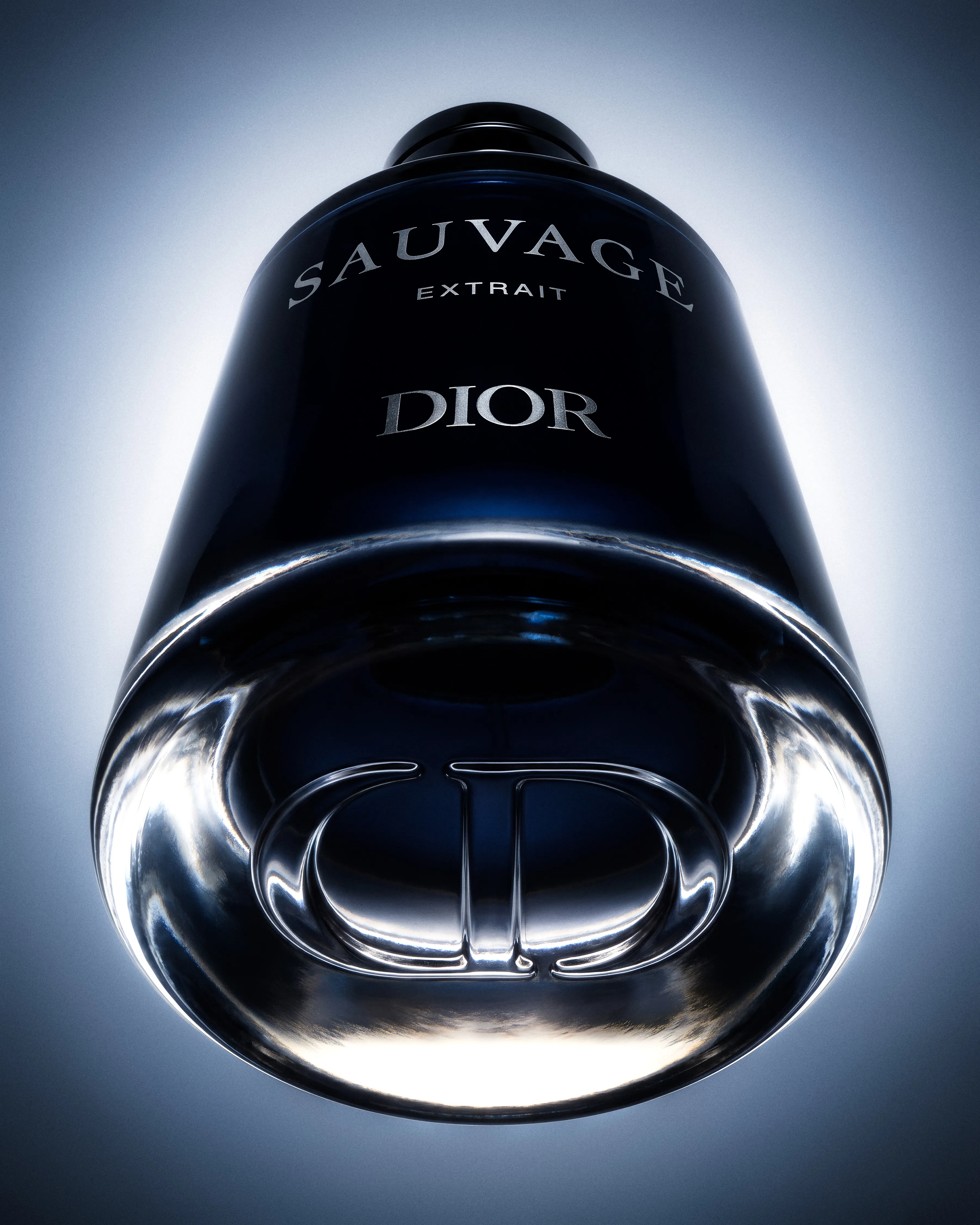 Extreme low-angle photograph of the Dior Sauvage Extrait bottle, enlarging the engraved CD monogram in its clear glass base against cold blue-white light.