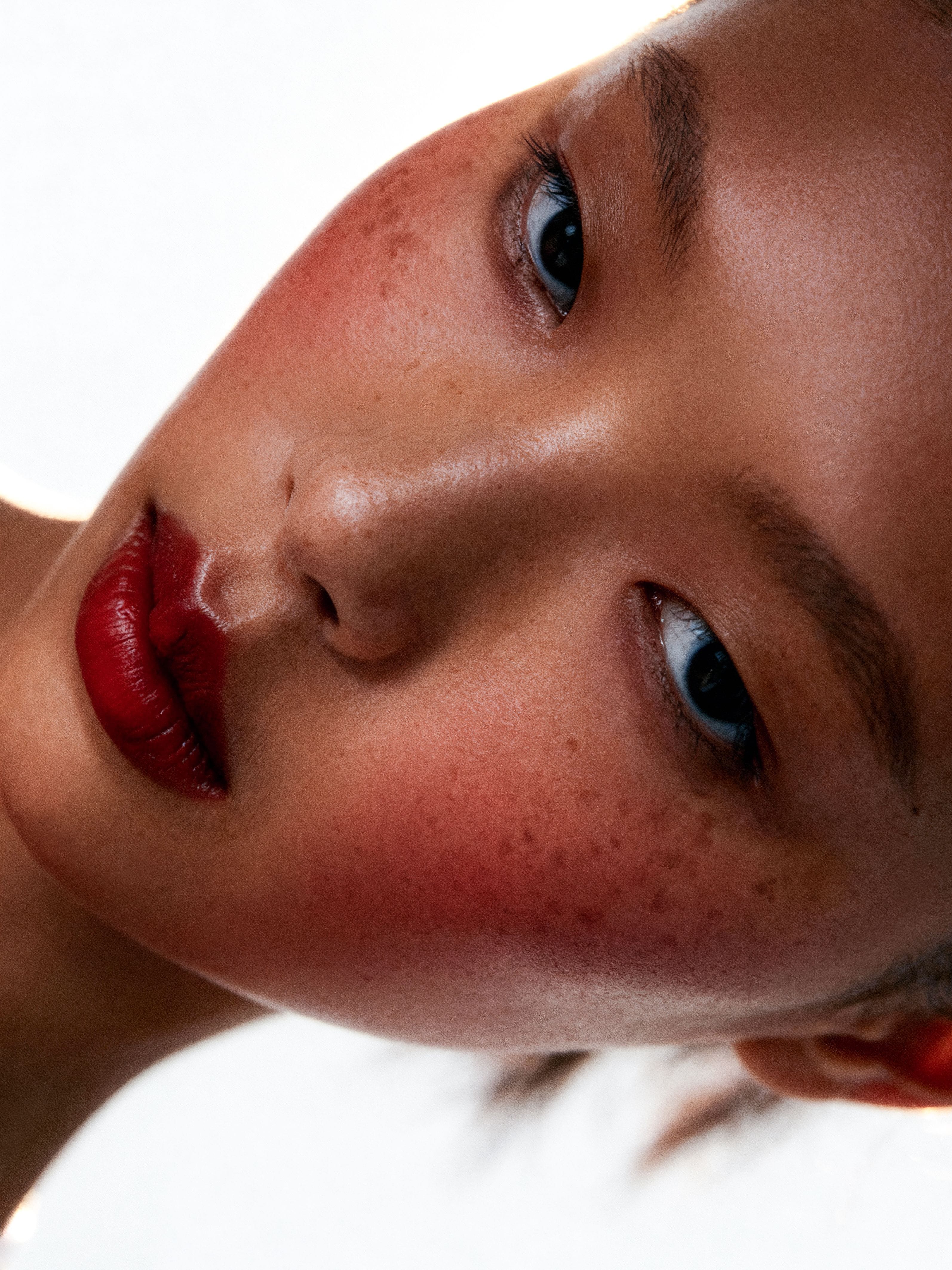 Rotated close beauty portrait with freckles, saturated red lips and a small teal accent at the inner eye.