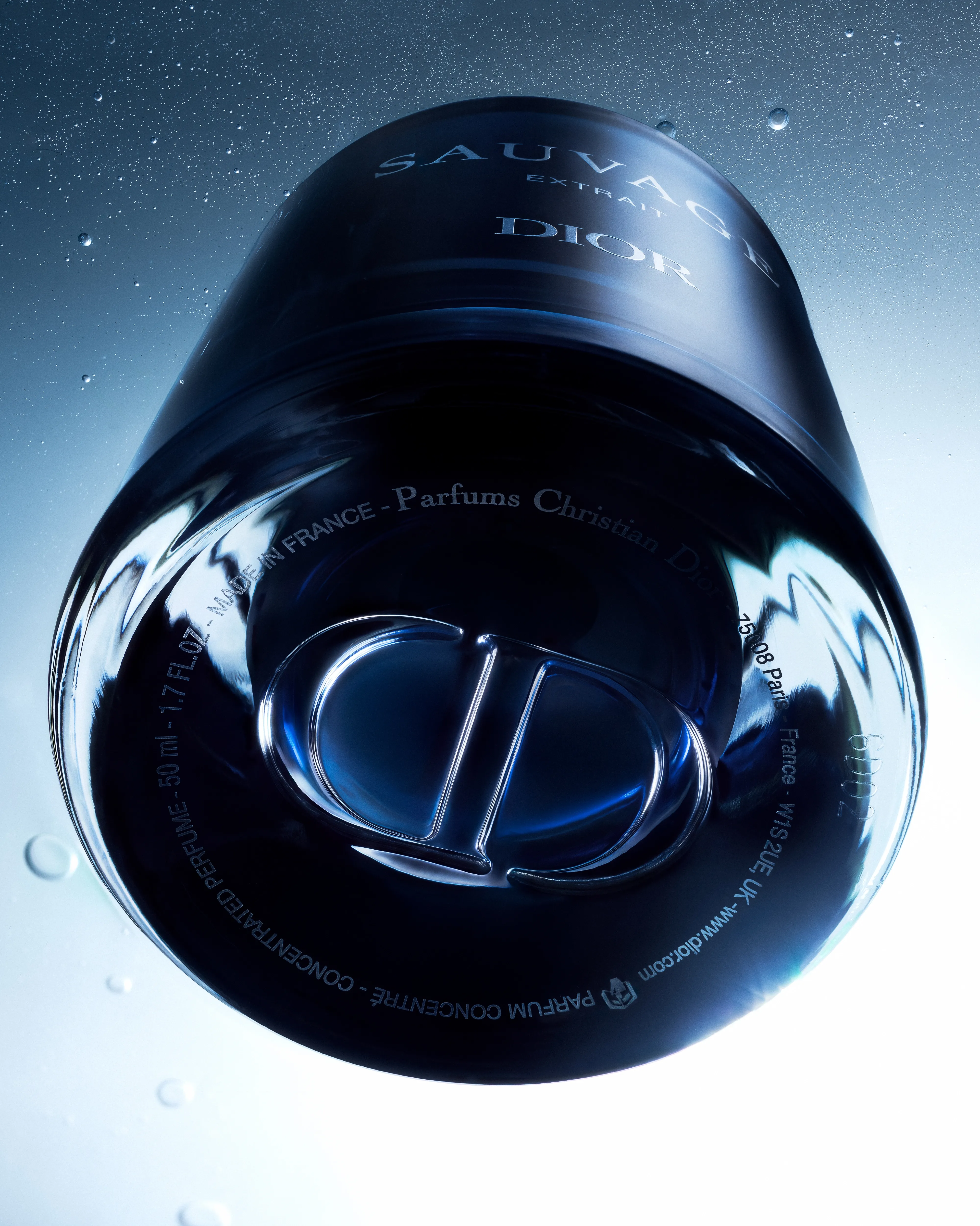 Extreme underside view of the Dior Sauvage Extrait bottle, centring the engraved CD monogram and circular base text amid cool light and fine droplets.