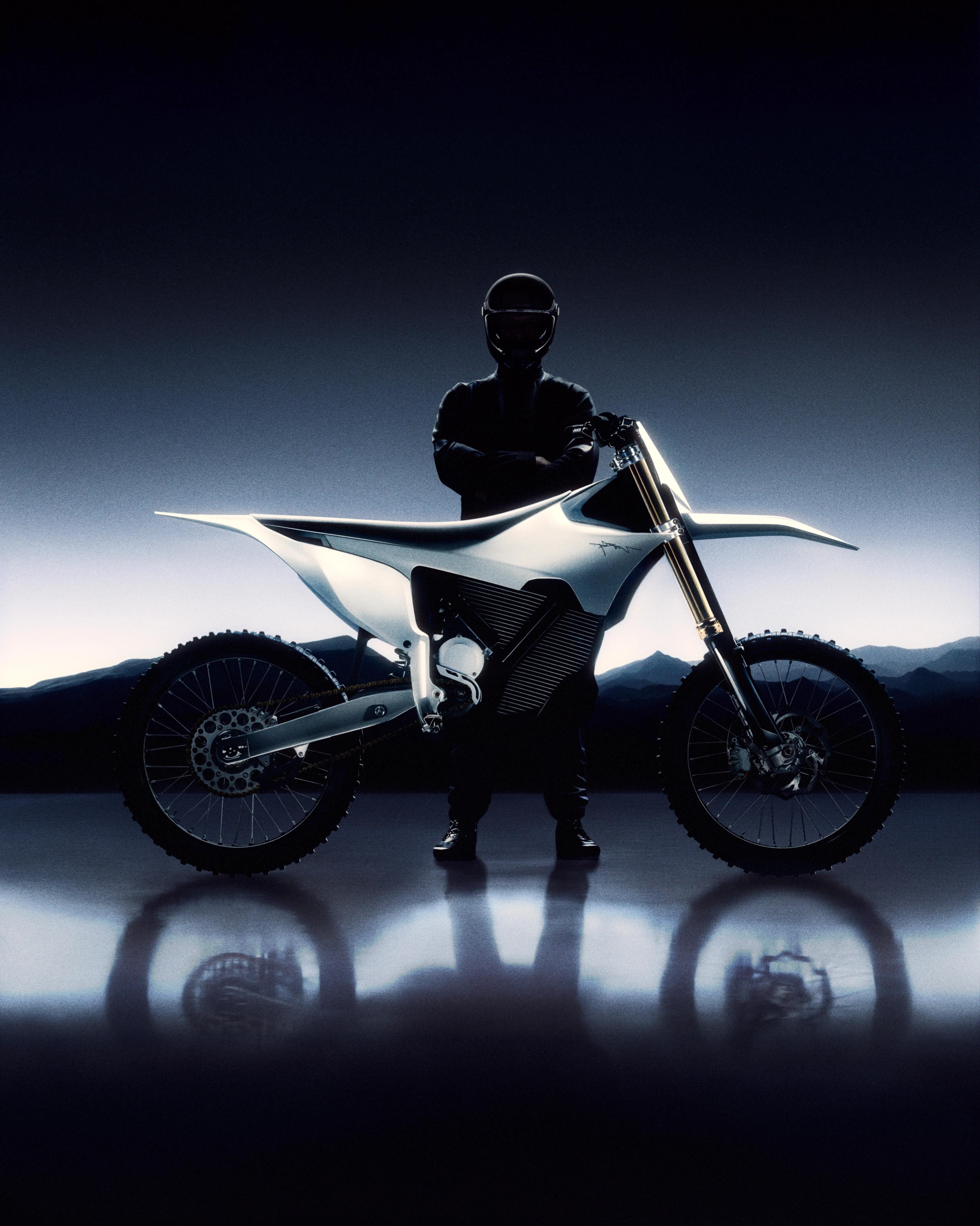 Side profile of a white Stark VARG electric motocross bike with a helmeted rider standing behind it in a dark studio.