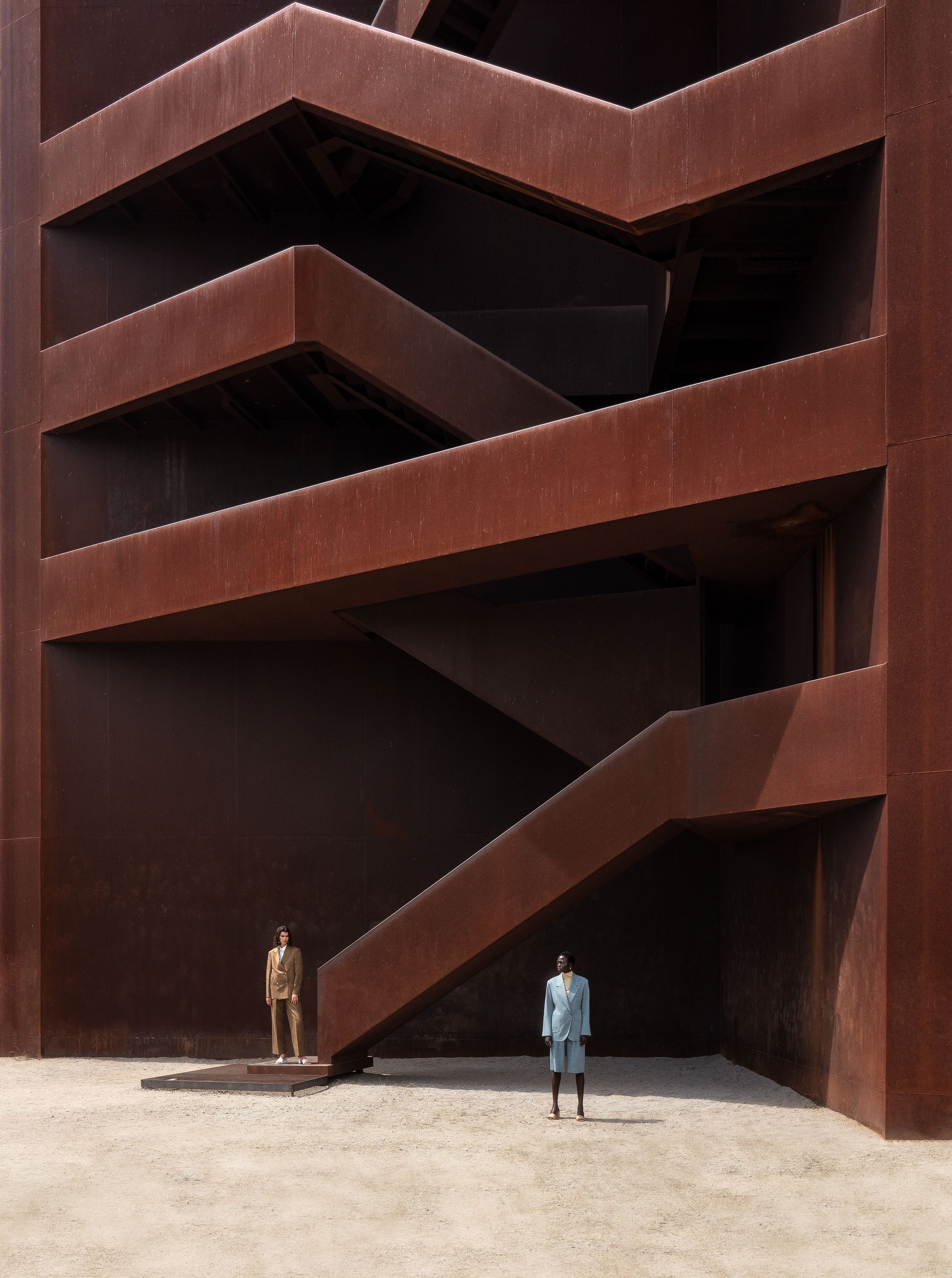 Two models standing at the base of a monumental rust-red structure crossed by angular stairways.